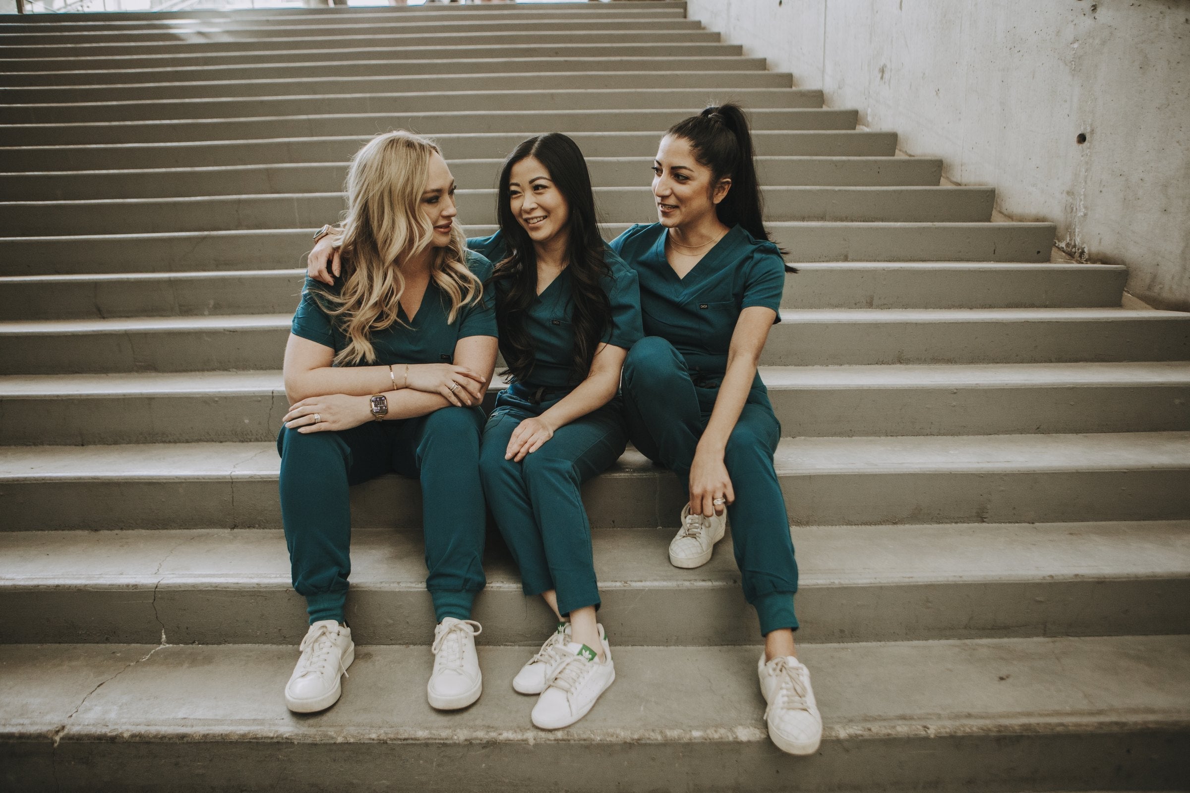 Three healthcare workers in teal KOA scrub sets sitting together on concrete stairs