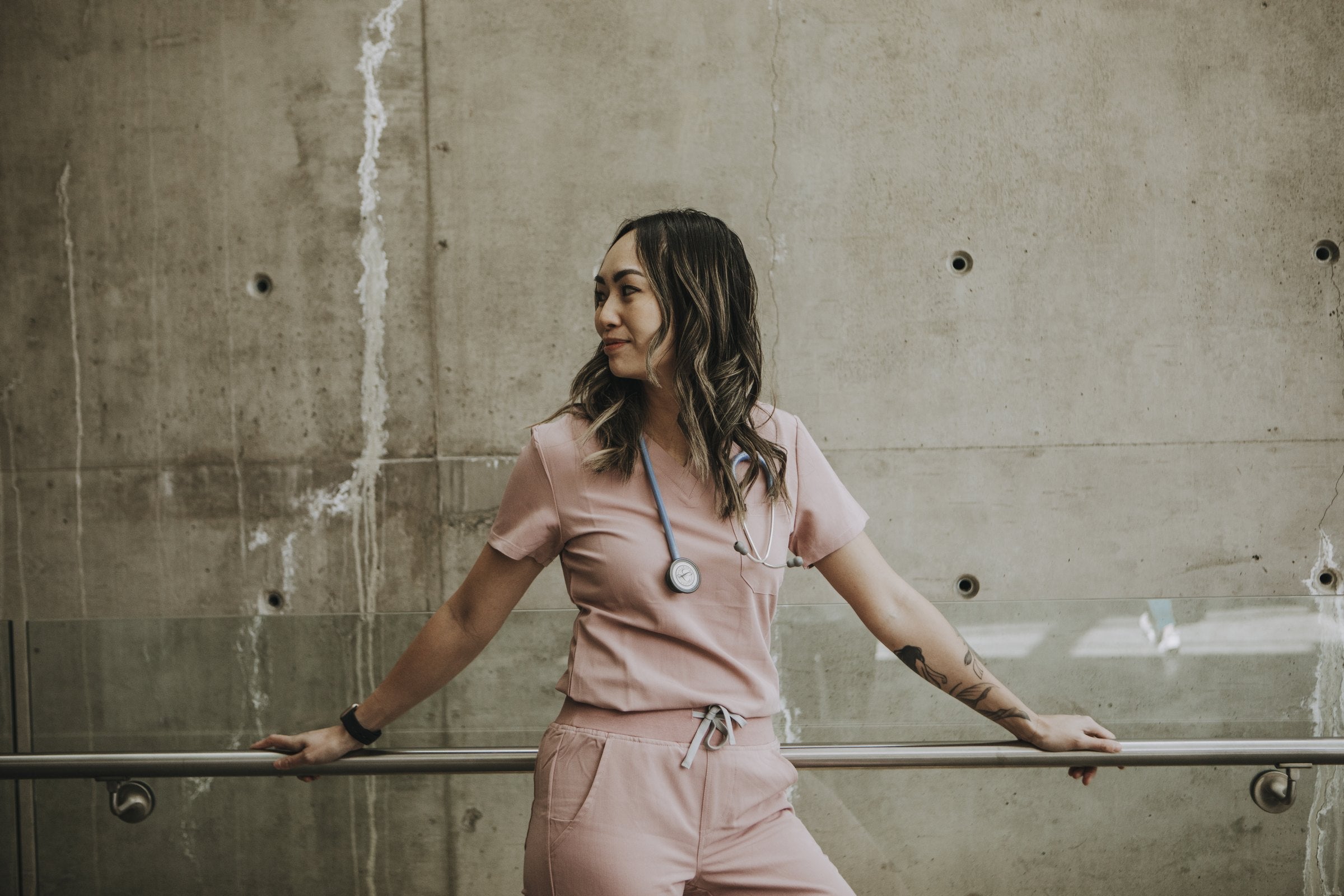 Healthcare worker in a blush KOA scrub set leaning against a concrete wall