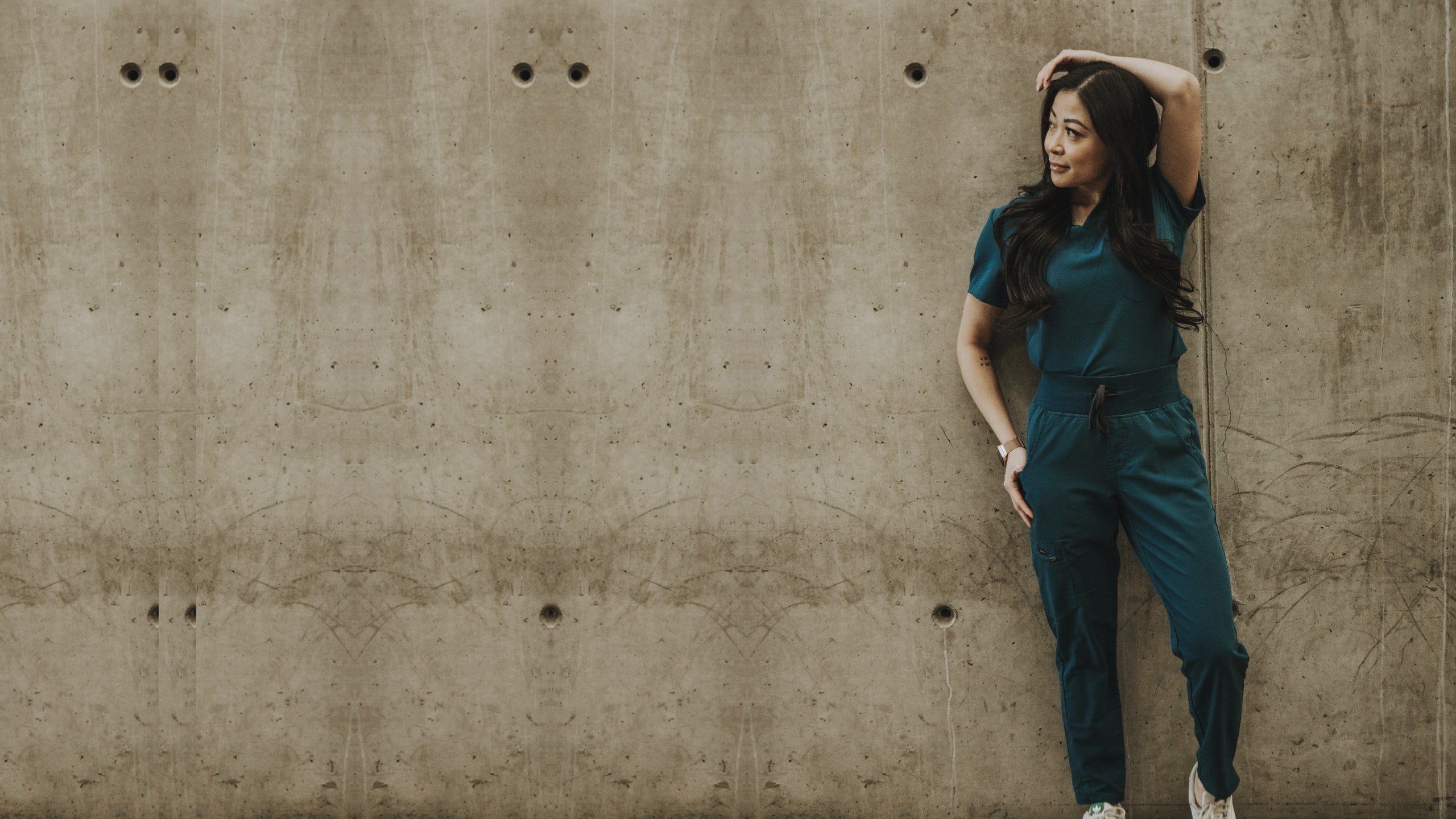Woman in a deep teal KOA scrub set - V-neck top and joggers with contrast drawstring - leaning on a concrete wall